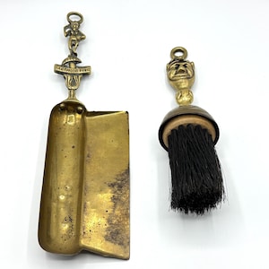 Può includere: Un set da camino in ottone con una paletta e una spazzola. La paletta ha un manico con una piccola figurina di un folletto e il testo "Dartmoor Pixie". La spazzola ha un manico con una piccola figurina di testa di diavolo.