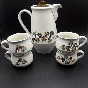 Puede incluir: Una tetera de cerámica blanca y cuatro tazas de cerámica blanca con un patrón de trébol verde. La tetera tiene una tapa marrón y las tazas tienen un borde marrón.