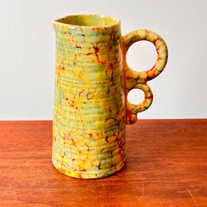 Puede incluir: Jarra alta de cerámica con un diseño de asa único. La jarra es principalmente amarilla con una superficie texturizada y detalles en rojo y verde. El asa presenta dos aberturas circulares. Probablemente se utiliza para servir bebidas.