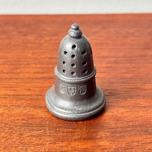 May include: A small, antique pewter salt shaker with a bell shape. The shaker has a series of small holes near the top and decorative emblems near the base. The salt shaker is sitting on a wooden surface.