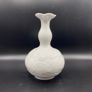 Puede incluir: Jarrón de cerámica blanco con un borde festoneado y un cuerpo bulboso texturizado. El jarrón tiene un diseño delicado y elegante.