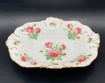 Może przedstawiać: Biała porcelana serwująca miska ze złotą obwódką i kwiatowym wzorem z różowymi różami. Miska ma falisty brzeg i lekko uniesiony środek.