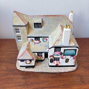 May include: A miniature model of a two-storey building with white walls, a textured grey roof, and black window frames. The exterior features colourful floral accents, two chimneys, and a sign that reads The Fisherman's Rest Circa 1302.