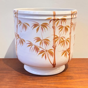 Peut inclure: Un pot de fleurs en céramique blanche de forme cylindrique et large, avec une base en piédestal. Le pot est décoré d'un motif répété de tiges et de feuilles de bambou dans des tons de marron et d'orange. Le pot est posé sur une surface en bois.