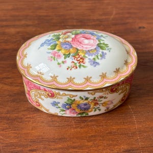 May include: An oval porcelain trinket box featuring a white base with a colorful floral bouquet on the lid. The edges are decorated with a scalloped pink and gold trim, while the sides display intricate pink and gold scrollwork and floral patterns.