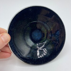May include: A small, dark blue ceramic dish with a glossy finish. The dish has a slight inward curve and a raised centre.