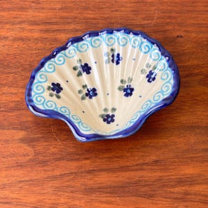 Puede incluir: Un plato de cerámica con forma de concha que presenta una base de color crema con motivos florales azul oscuro y hojas verdes pintados a mano. El borde está decorado con un diseño de espirales azul claro y un borde festoneado azul oscuro.