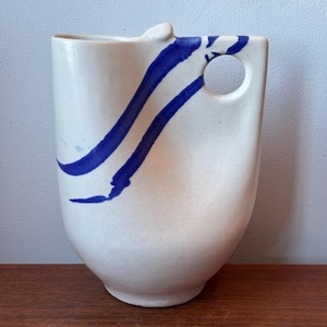 Puede incluir: Una jarra de cerámica de color crema con acabado moteado y un diseño de pincelada azul. El recipiente presenta una silueta única con un asa circular recortada integrada en la parte superior derecha y un pequeño pico vertedor.