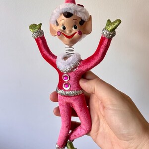 Könnte beinhalten: Vintage Weihnachts-Elf-Ornament mit Wackelkopf-Design. Der Elf trägt ein rosa Samt-Outfit mit silbernen Glitzerakzenten. Das Gesicht des Elfen hat rosige Wangen und einen lächelnden Ausdruck. Der Hut des Elfen ist rosa mit weißem Besatz.