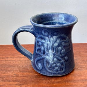 Könnte beinhalten: Eine blaue Keramiktasse mit Henkel. Die Tasse hat ein erhabenes Blumendesign und eine dunkelblaue Glasur. Die Tasse steht auf einer Holzoberfläche.