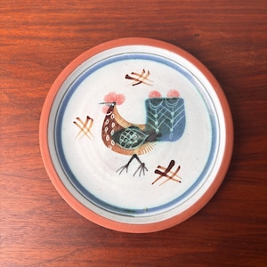 Puede incluir: Un plato de cerámica con un diseño de gallo. El plato tiene un centro blanco con un borde azul y marrón. El gallo es marrón, azul y naranja con una cresta roja. El plato tiene un borde marrón.