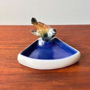 Puede incluir: Un cuenco de cerámica con interior azul y borde blanco, coronado por una detallada figura de pájaro. El pájaro tiene plumas marrones, negras y grises. El cuenco es triangular y está sobre una superficie de madera.