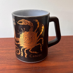May include: Black ceramic mug with a gold crab zodiac design. The mug features the word "Cancer" and the phrase "the crab". The handle is black, and the interior is light blue.