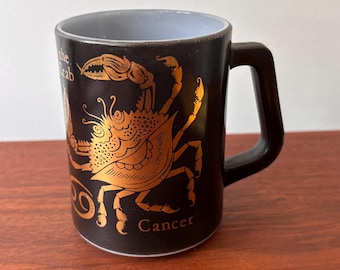 Pode incluir: Caneca de cerâmica preta com um design dourado do signo do zodíaco de Câncer. A caneca apresenta a palavra "Cancer" e a frase "the crab". A alça é preta e o interior é azul claro.