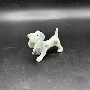 May include: A white porcelain figurine of a dog with blue and gray markings. The dog is standing on all four legs with its head tilted up and its mouth open.