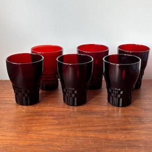 Op de afbeelding: Een set van zes dieprode glazen bekers gerangschikt in twee rijen. Elke beker heeft een gladde bovenrand en een getextureerde basis met een herhalend vierkant rasterpatroon. Het glas is doorschijnend en laat licht door de levendige rode kleur.