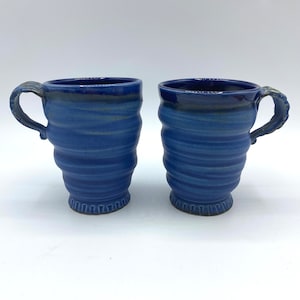 May include: Two blue ceramic mugs with a wavy design. The mugs have a handle on the right side.