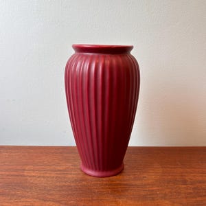 May include: A burgundy-colored vase with vertical ribbed detailing. The vase has a rounded base and a slightly flared top. The vase is made of a matte material and sits on a wooden surface.