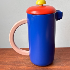 May include: A colorful coffee press with a blue body, red lid, yellow knob, and a pink handle. The coffee maker is cylindrical and sits on a wooden surface. The design is modern and eye-catching.