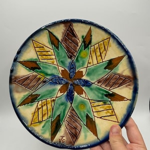 Peut inclure: Une assiette en céramique avec un bord bleu et un motif géométrique coloré. Le motif présente un motif en étoile avec des couleurs vertes, jaunes, brunes et bleues.