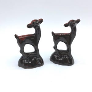 May include: Two ceramic deer figurines. The deer are dark brown with a reddish-brown interior. Each deer has an open space in the middle of its body and stands on a small base. The figurines are on a white background.