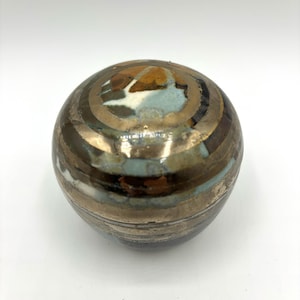 May include: A small, round ceramic trinket box with a multi-colored glaze. The box has a dark brown base with a light blue and white center. The top of the box is decorated with gold and brown stripes.