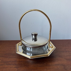 May include: A clear glass serving dish with a silver-toned lid and a central knob, set within a hexagonal silver and gold-toned metal base. A tall, arched gold-toned metal handle is attached to the base, extending vertically over the lid.