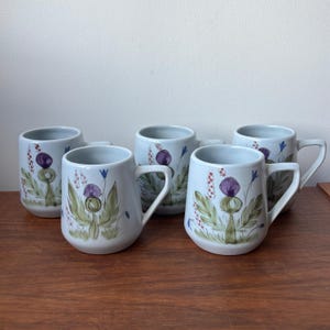 May include: A set of five white ceramic mugs, each featuring a hand-painted botanical design of a purple thistle flower with green leaves and small red and blue accents. The mugs have a rounded body shape, a smooth finish, and a sturdy side handle.