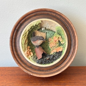 May include: A circular wall plaque featuring a three-dimensional relief of a small brown and grey bird perched on a terracotta-coloured pipe. The scene includes green foliage, orange flowers, and a wooden fence, all encased in a dark wood frame.