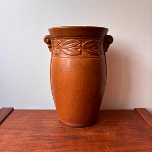 Könnte beinhalten: Eine braune Keramikvase mit einem dekorativen Band aus Blättern und Punkten am oberen Rand. Die Vase hat zwei kleine Griffe und eine glatte, zylindrische Form. Die Vase steht auf einer Holzoberfläche.