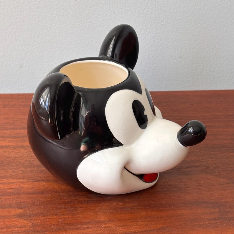 May include: A ceramic Mickey Mouse head-shaped mug. The mug features a black and white design with a red mouth and a black nose. The mug is sitting on a wooden surface.