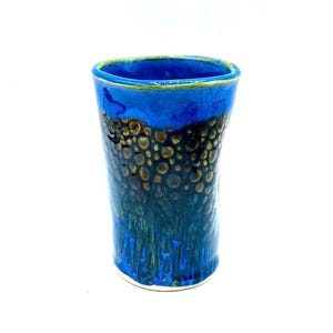May include: A handmade ceramic vase with a vibrant blue and gold speckled design. The vase has a cylindrical shape with a slightly flared top and a textured surface. The rim is accented with a green glaze.
