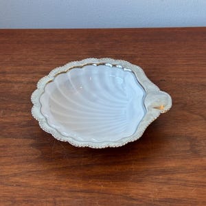 May include: A scallop shell-shaped dish featuring a white, translucent centre with ribbed detailing and a metallic silver-toned rim. The edge of the dish has a scalloped, beaded texture, providing a decorative border around the central basin.