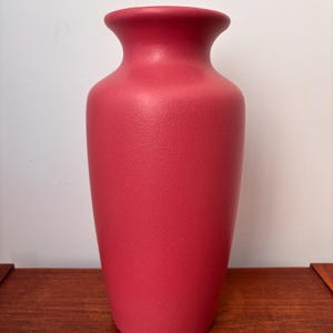 May include: A solid red vase with a textured surface. The vase has a wide body that tapers to a narrow neck and flared rim. The vase is sitting on a wooden surface.
