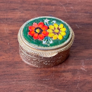 May include: A small, round, gold-toned trinket box with a mosaic floral design on the lid. The mosaic features red and yellow flowers set against a green background. The box has an ornate, engraved pattern around the sides.