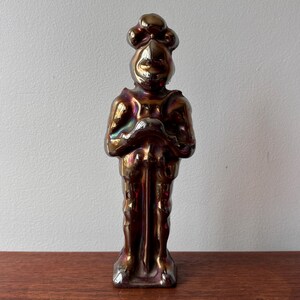 Może przedstawiać: Ceramiczna figurka rycerza z mieczem, stojąca na drewnianej powierzchni. Rycerz jest w zbroi i ma hełm z pióropuszem. Figurka jest tęczowa w odcieniach fioletu, złota i brązu.