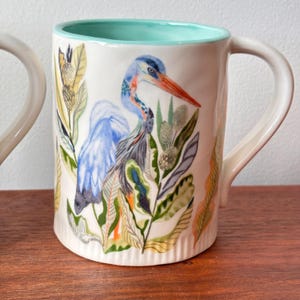 Puede incluir: Taza de cer&aacute;mica blanca con interior turquesa y asa grande. La taza presenta una ilustraci&oacute;n detallada de una garza azul rodeada de follaje colorido. La garza tiene un largo pico naranja sobre un fondo blanco.