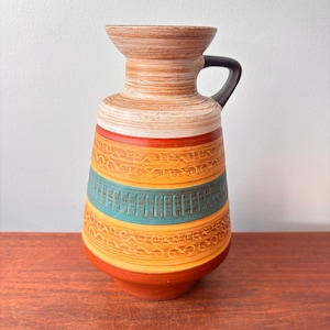 May include: A vintage ceramic vase with a handle, featuring a color palette of orange, yellow, and teal. The vase has a textured surface with a patterned design and a dark handle. The vase is a decorative item.
