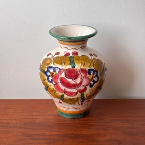 May include: A hand-painted ceramic vase with a cream-colored base, featuring a large red rose surrounded by green and gold leaves. The vase has a green rim and base, and is displayed on a wooden surface.