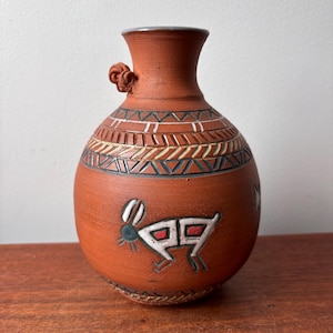 May include: Terracotta ceramic vase with a rounded body and a narrow neck. The vase features a hand-painted design of a rabbit and geometric patterns in white, red, and blue. A small handle is on the side. The vase is approximately 20 cm tall.