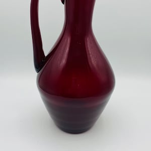 May include: A deep red glass jug with a rounded body and a curved handle.
