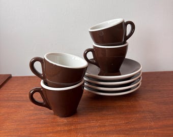 Könnte beinhalten: Ein Set aus Espressotassen und Untertassen. Die Tassen sind dunkelbraun mit weißen Innenräumen und Rändern. Die Untertassen sind dunkelbraun mit weißen Rändern. Die Tassen sind gestapelt und die Untertassen liegen in einem ordentlichen Stapel. Das Set steht auf einer Holzoberfläche.