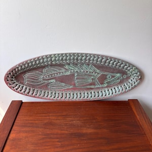 Puede incluir: Una fuente ovalada de cerámica con un diseño de pez. La fuente es de color marrón rojizo con un pez y un borde verdes. El pez está detallado con aletas, escamas y una aleta dorsal. El borde tiene un patrón repetitivo.