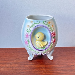 May include: A pastel blue and white ceramic egg-shaped planter with a yellow chick inside a purple-bordered oval. The planter is decorated with pink and yellow flowers and green vines, and has three white feet.