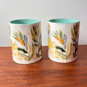 Puede incluir: Dos tazas cil&iacute;ndricas de cer&aacute;mica con base color crema y borde verde azulado claro. Las tazas est&aacute;n decoradas con un colorido estampado floral con elementos bot&aacute;nicos amarillos, naranjas, verdes y azules. Las tazas se exhiben sobre una superficie de madera.