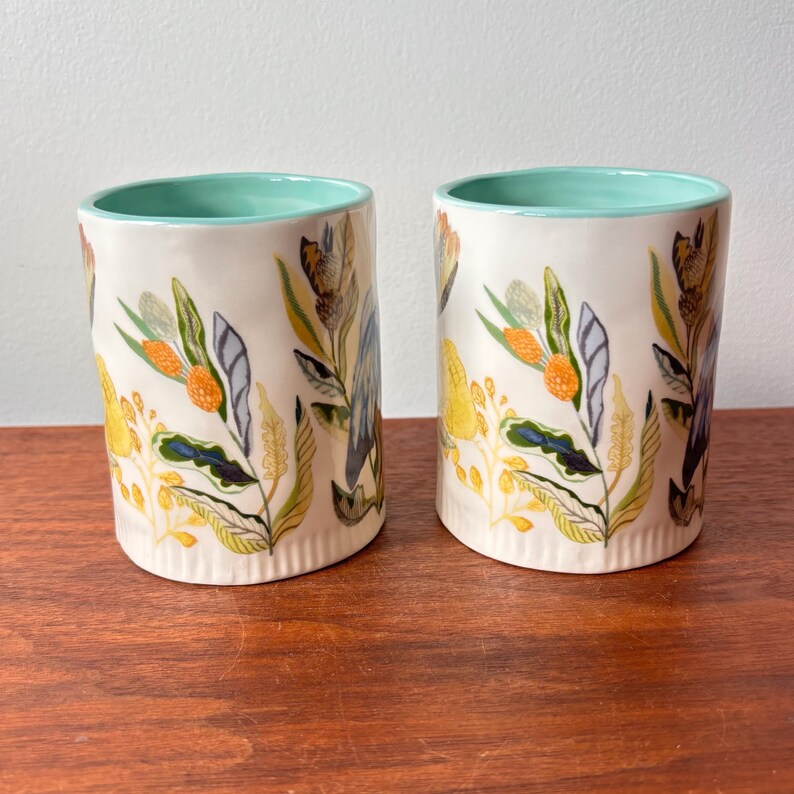 Puede incluir: Dos tazas cil&iacute;ndricas de cer&aacute;mica con base color crema y borde verde azulado claro. Las tazas est&aacute;n decoradas con un colorido estampado floral con elementos bot&aacute;nicos amarillos, naranjas, verdes y azules. Las tazas se exhiben sobre una superficie de madera.
