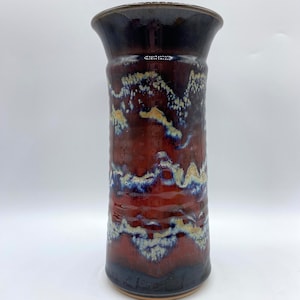 May include: A tall, cylindrical ceramic vase with a dark blue glaze and a red and white abstract pattern. The vase has a slightly flared opening.