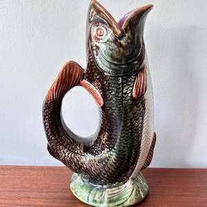 May include: A ceramic fish-shaped jug, predominantly brown, green, and white. It features a brown handle and tail, with the mouth open. The base is green with leaf detailing. The jug is a decorative item.