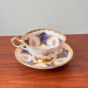 Op de afbeelding: Een vintage theekop en schotel set. De witte porseleinen kop en schotel hebben een gouden rand, blauwe en gouden sierlijke ontwerpen en kleine roze bloemaccenten. De kop heeft een gebogen handvat.