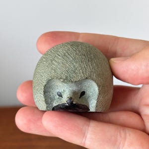 Pode incluir: Uma pequena escultura de pedra cinzenta em forma de ouriço, com uma superfície texturizada e cruzada. A figura apresenta dois pequenos olhos pintados de preto e um nariz e boca pintados de castanho escuro, criando uma representação animal compacta.